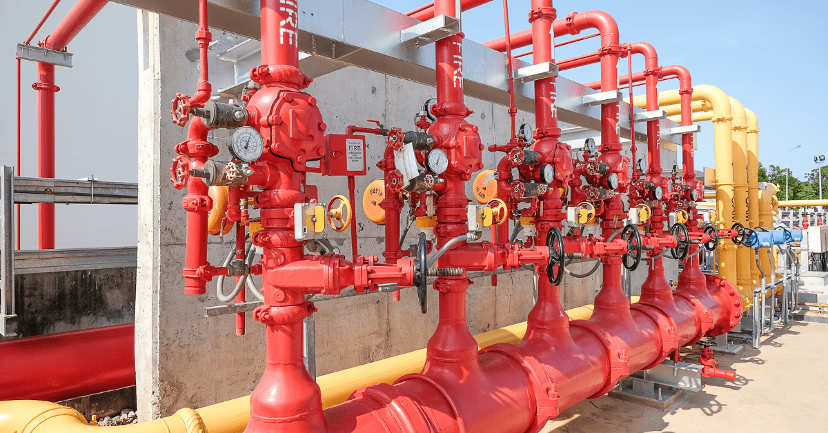 A row of red fire hydrants and valves connected by yellow pipes, with gauges and control panels mounted on a concrete wall. Fire Alarm Surge Protection is a crucial component of this fire suppression system, ensuring its reliability and preventing disruptions during emergencies.